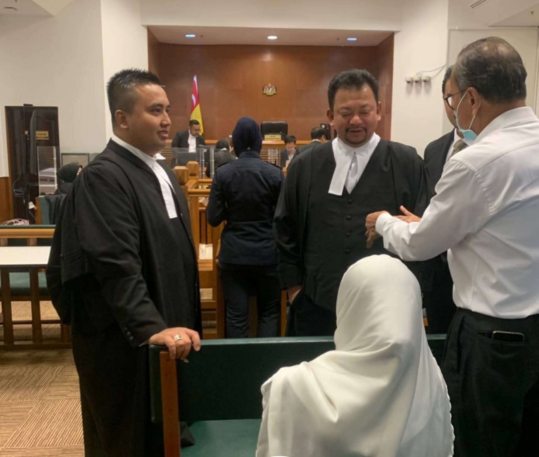 Two advocates confer in a courtroom beneath the Malaysian coat of arms.
