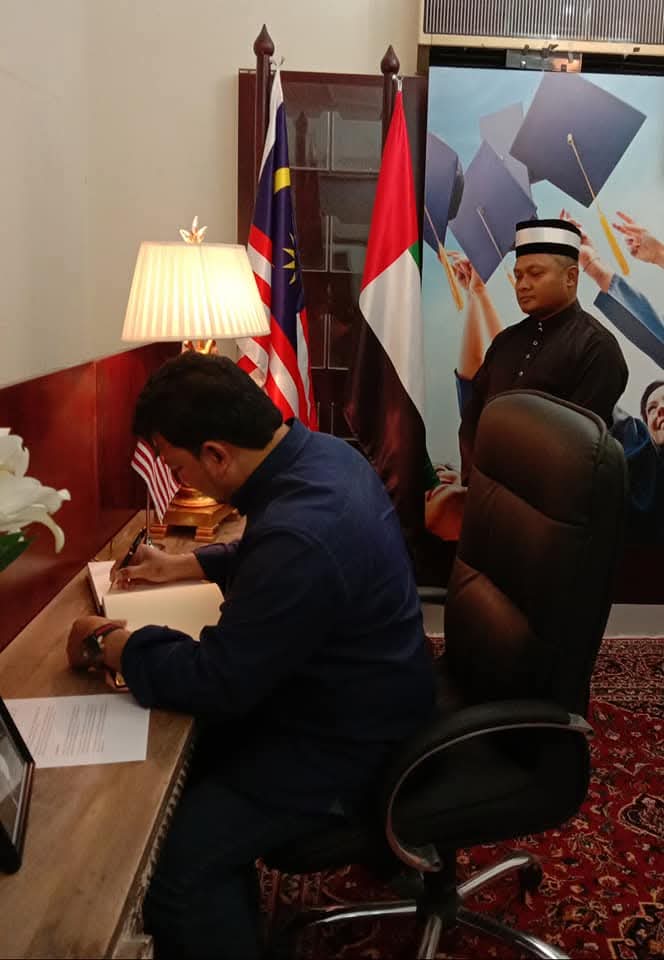 Signing a book at a desk flanked by the Malaysian and UAE flags.