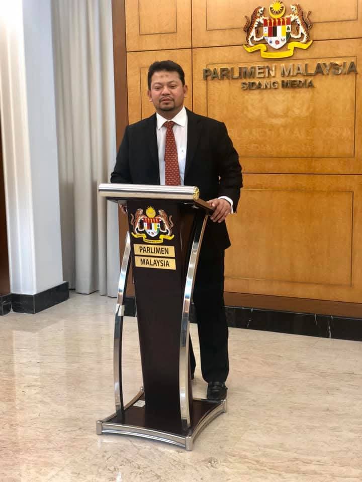 A member of the firm speaking at a press conference lectern at the Malaysian Parliament.